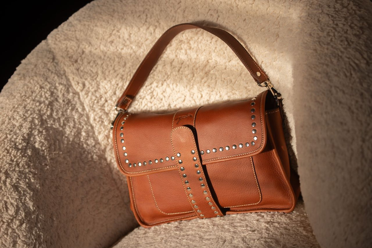 Trois sacs Pénélope en cuir cognac — un cabas, un sac à bandoulière tissée et un sac à rabat clouté — posés sur une cheminée en marbre devant des boiseries dorées.